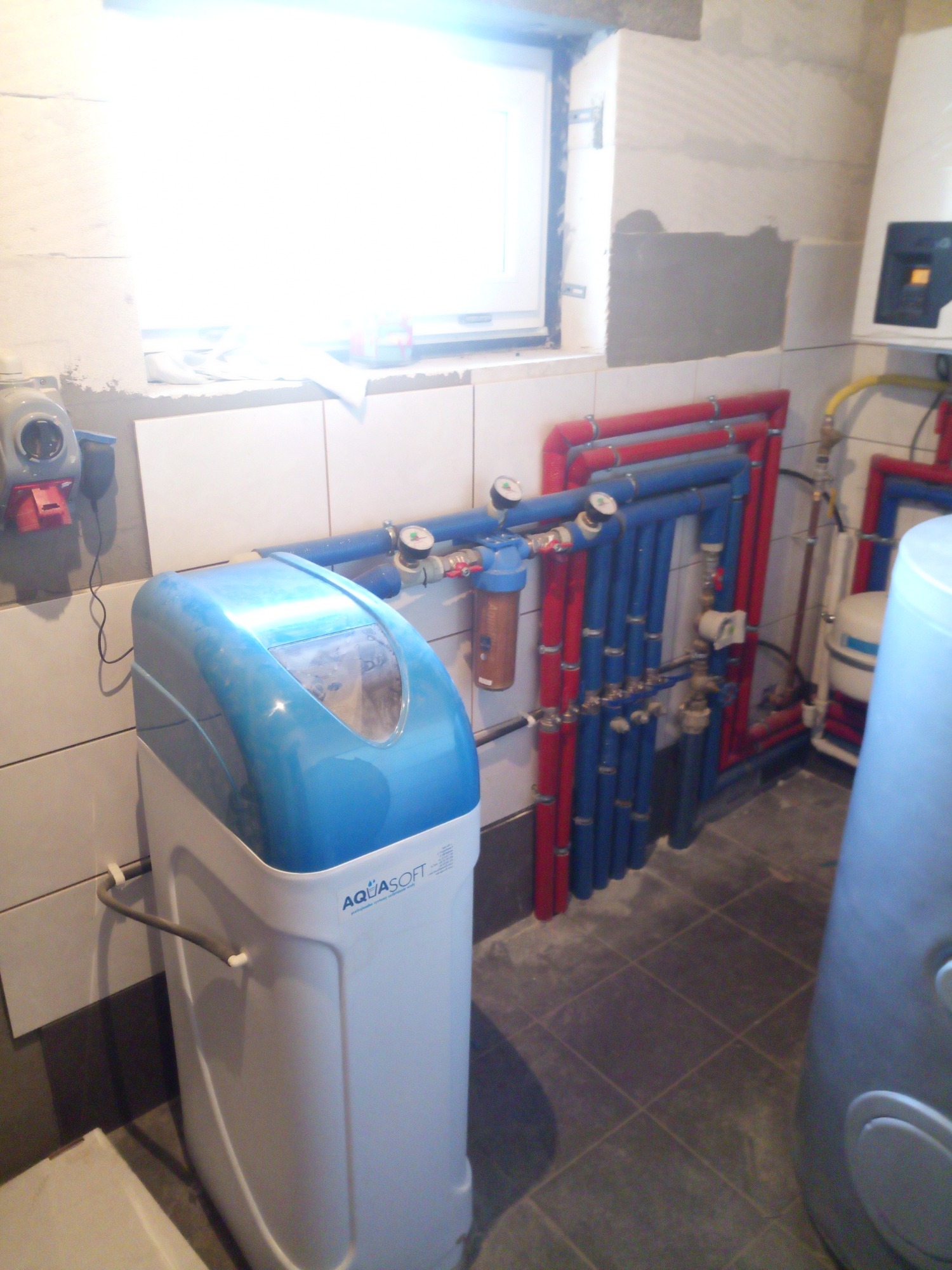 Biało-błękitny zmiękczacz wody AquaSoft zainstalowany w pomieszczeniu z płytkami, widoczne rury instalacji wodnej w kolorze czerwonym i niebieskim, oraz okno w tle.