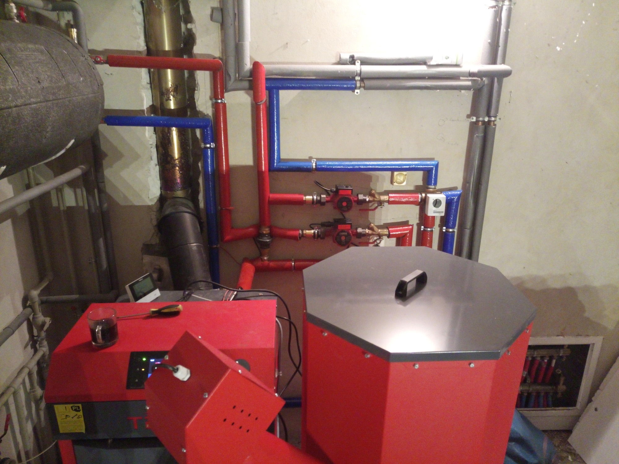 Czerwony piec z podajnikiem na pellet, widoczne rury instalacji grzewczej w kolorach czerwonym i niebieskim, na piecu stoi szklanka z ciemnym płynem oraz wkrętak.