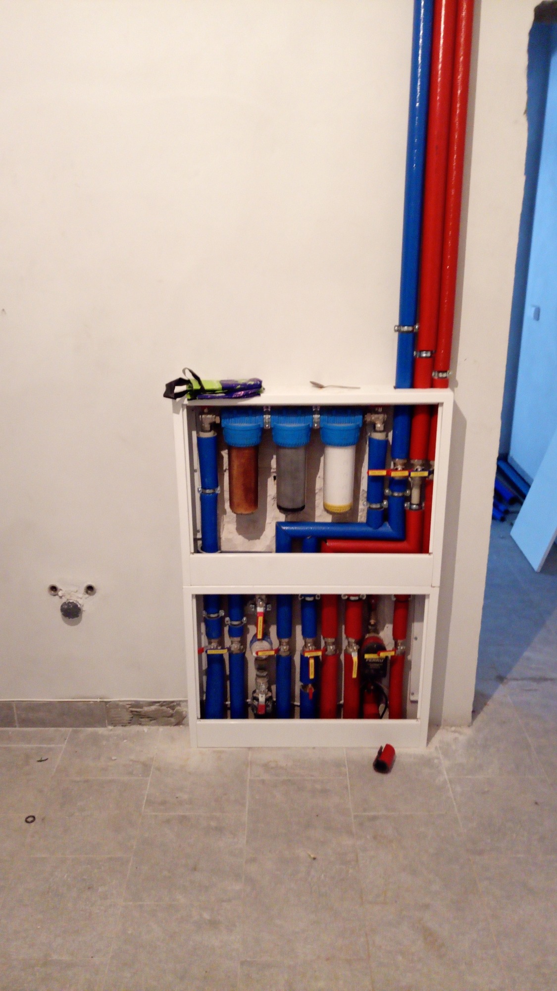 Instalacja filtrów wody w obudowie z rurami doprowadzającymi w kolorze czerwonym i niebieskim, widok na ścianę z dwoma otworami, Czerwin.