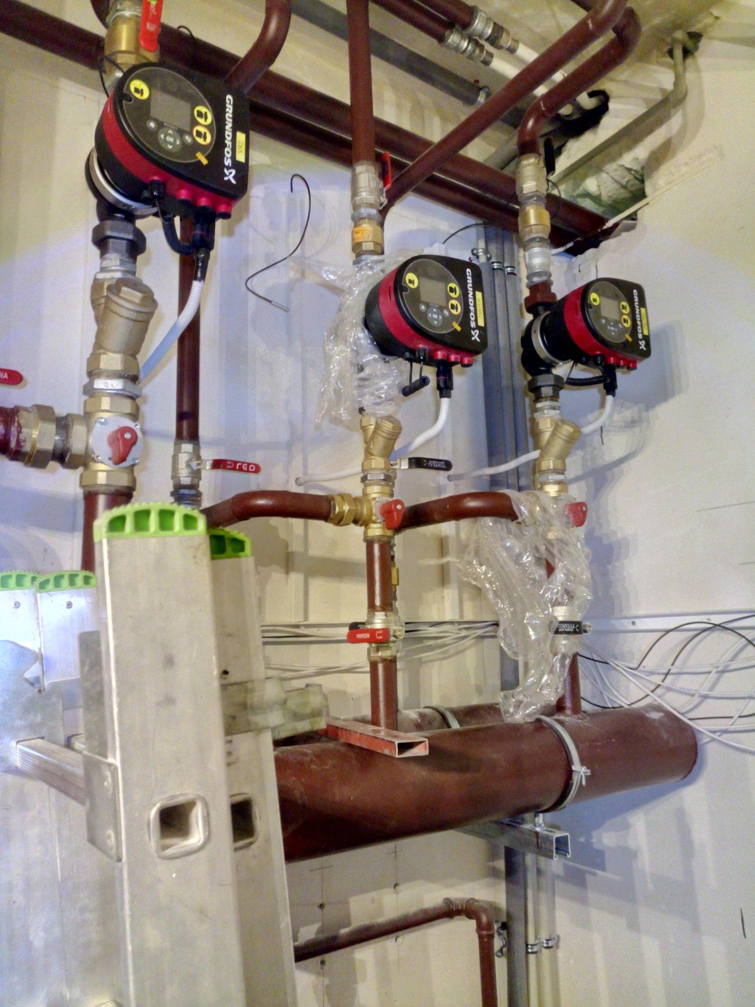 Instalacja centralnego ogrzewania z trzema pompami obiegowymi Grundfos, rurami miedzianymi i zaworami, widoczna drabina aluminiowa z zielonymi nakładkami na szczeblach.