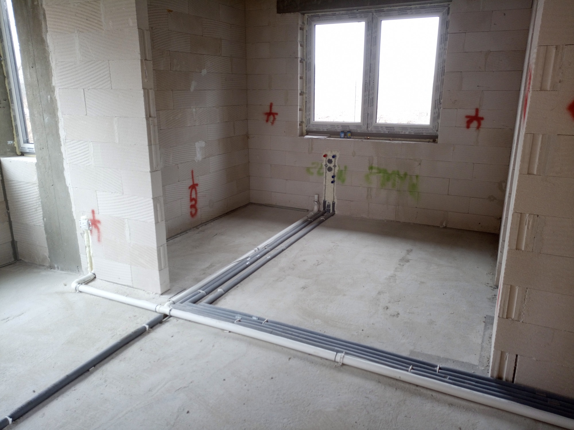Surowy stan wnętrza z widocznymi rurami instalacji wodnej i kanalizacyjnej na podłodze, ściany z betonu komórkowego z oznaczeniami instalacyjnymi w kolorze czerwonym, okno z białymi ramami w tle.