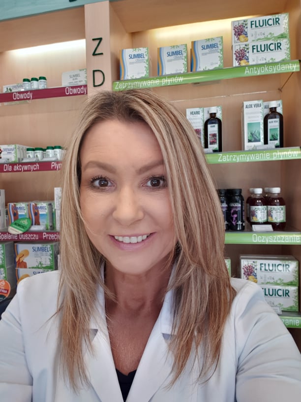 Uśmiechnięta kobieta w białym fartuchu na tle półek z suplementami diety, widoczne produkty wspomagające odchudzanie i detoks.