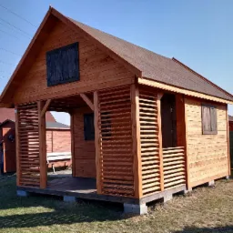 Drewniana altana z lamelami, podniesiona na betonowych bloczkach, z brązowym dachem i ciemnymi okiennicami, na tle trawnika i błękitnego nieba.