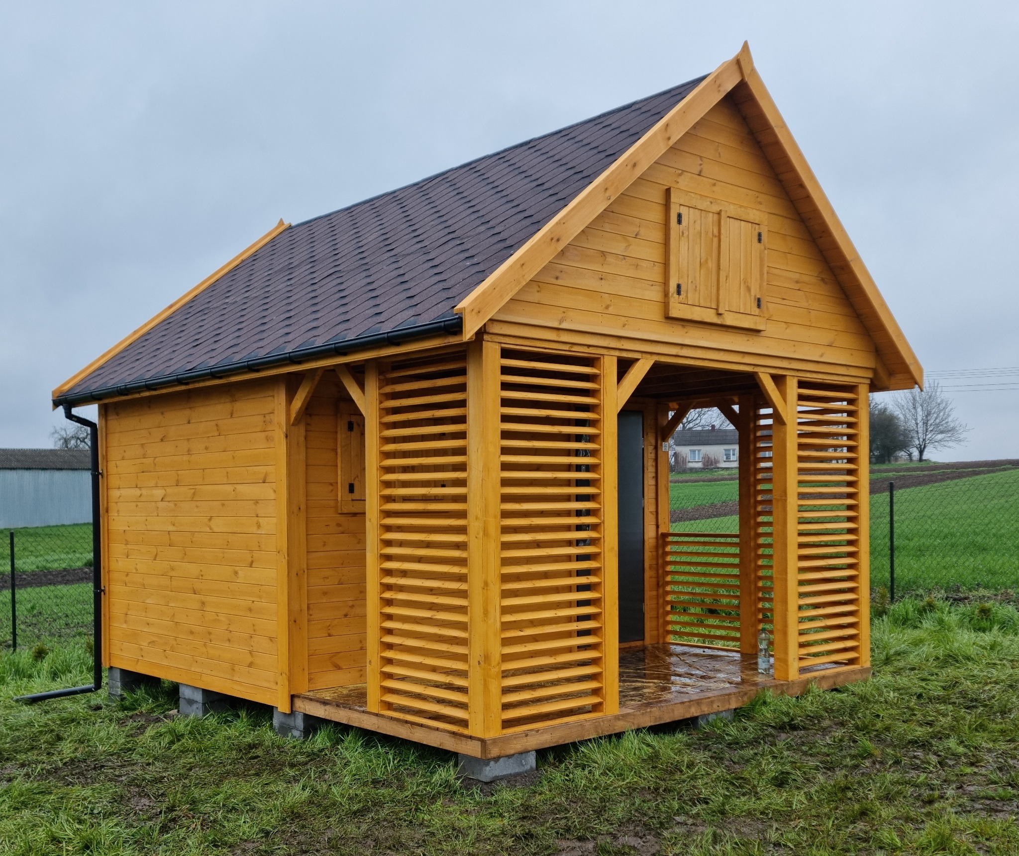Nowoczesna, drewniana altana z brązowym dachem bitumicznym, zbudowana na betonowych bloczkach, z lamelowymi ścianami i małymi okiennicami, na tle zielonego trawnika i pochmurnego nieba.