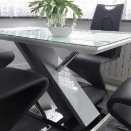 Szklany stół z geometryczną, szarą podstawą i czarnymi krzesłami o zakrzywionych oparciach, ustawiony na szarym dywanie, z widocznym grzejnikiem i firankami w tle.