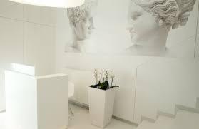 Jasne, minimalistyczne wnętrze recepcji kliniki z białymi meblami, donicą z kwiatami i reprodukcją klasycznych rzeźb na ścianie.