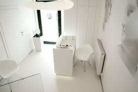 Jasne, minimalistyczne wnętrze gabinetu zabiegowego z białymi meblami, krzesłem i dekoracyjnym kwiatem w donicy. Na ścianie widoczny fragment grafiki przedstawiającej profil twarzy.