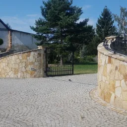 Wjazd na posesję wyłożony kostką brukową w odcieniach szarości, z ozdobnymi, łukowatymi murkami z kamienia naturalnego i czarną, kutą bramą w tle, częściowo wykończony budynek mieszkalny w tle.