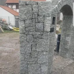 Surowa kolumna z kostki granitowej z widocznymi spoinami i konstrukcją łukową w tle, na tle budynków mieszkalnych i placu budowy.