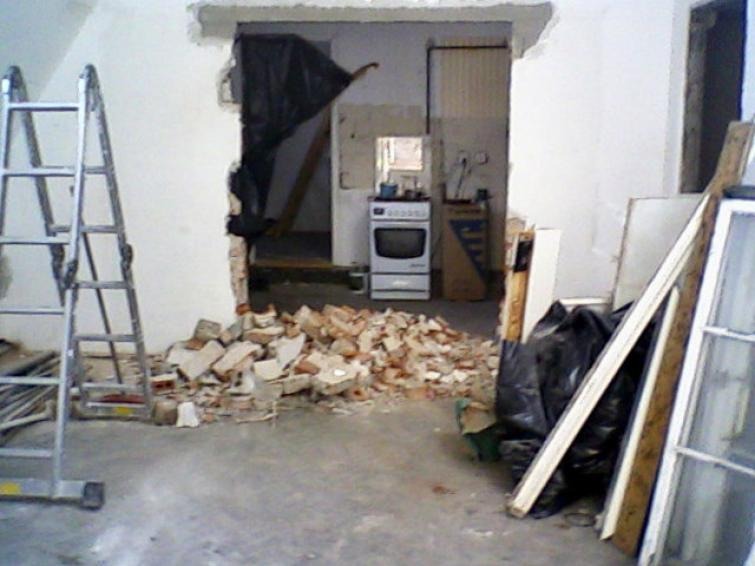Demontaż ściany działowej w mieszkaniu: sterta gruzu ceglanego na podłodze, widoczna drabina aluminiowa i wnętrze kuchni z kuchenką w tle, ściana częściowo osłonięta czarną folią zabezpieczającą.