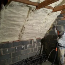 Pracownik w kombinezonie ochronnym aplikuje izolację na poddaszu z ceglanymi ścianami, widoczne drewniane belki stropowe.