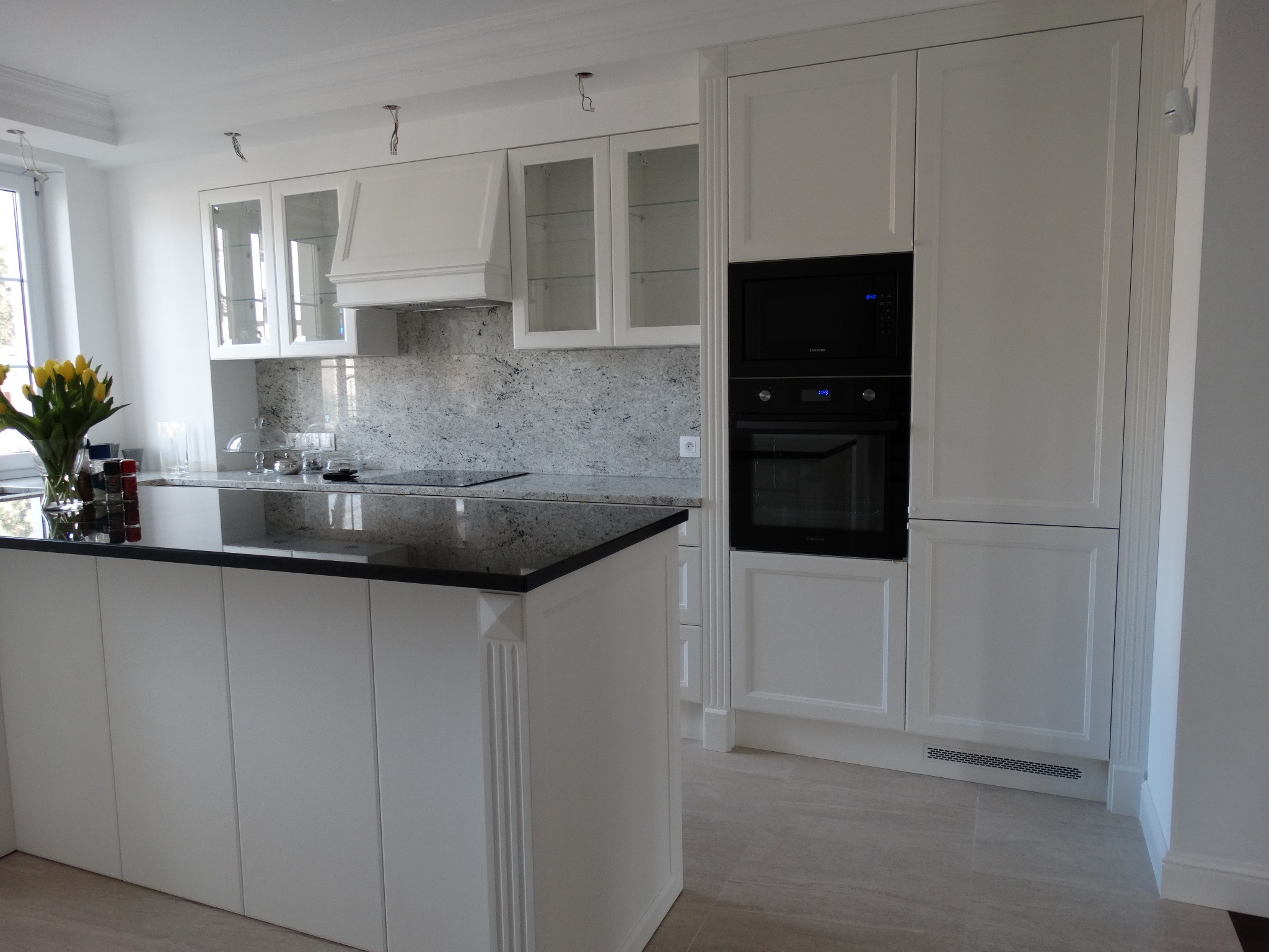 Biała kuchnia z wyspą, czarny blat, zabudowana piekarnikiem i szafkami z przeszklonymi frontami; w tle marmurkowy backsplash i bukiet żółtych tulipanów.