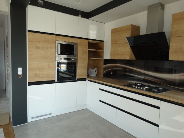 Nowoczesna kuchnia z białymi szafkami dolnymi z czarnymi uchwytami, szafkami górnymi z forniru drewnopodobnego, zabudowaną kuchenką mikrofalową i piekarnikiem, oraz szklanym panelem nad blatem...