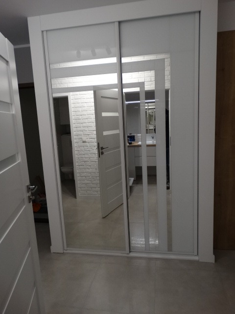 Biała szafa przesuwna z lustrzanymi drzwiami, odbijająca wnętrze łazienki z białą cegłą i toaletą, widok z boku.