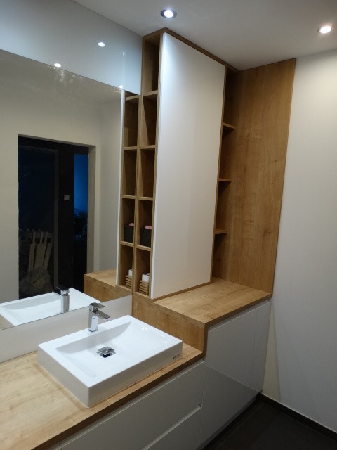 Nowoczesna łazienka z umywalką nablatową, drewnianym blatem i zabudową meblową z półkami, oświetleniem punktowym i dużym lustrem.