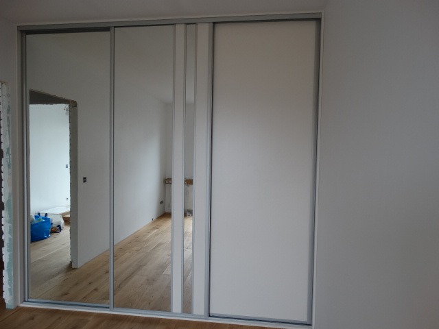 Wbudowana szafa z przesuwnymi drzwiami, część drzwi z lustrami, widok na wnętrze pokoju z drewnianą podłogą i elementami wykończeniowymi w trakcie prac.