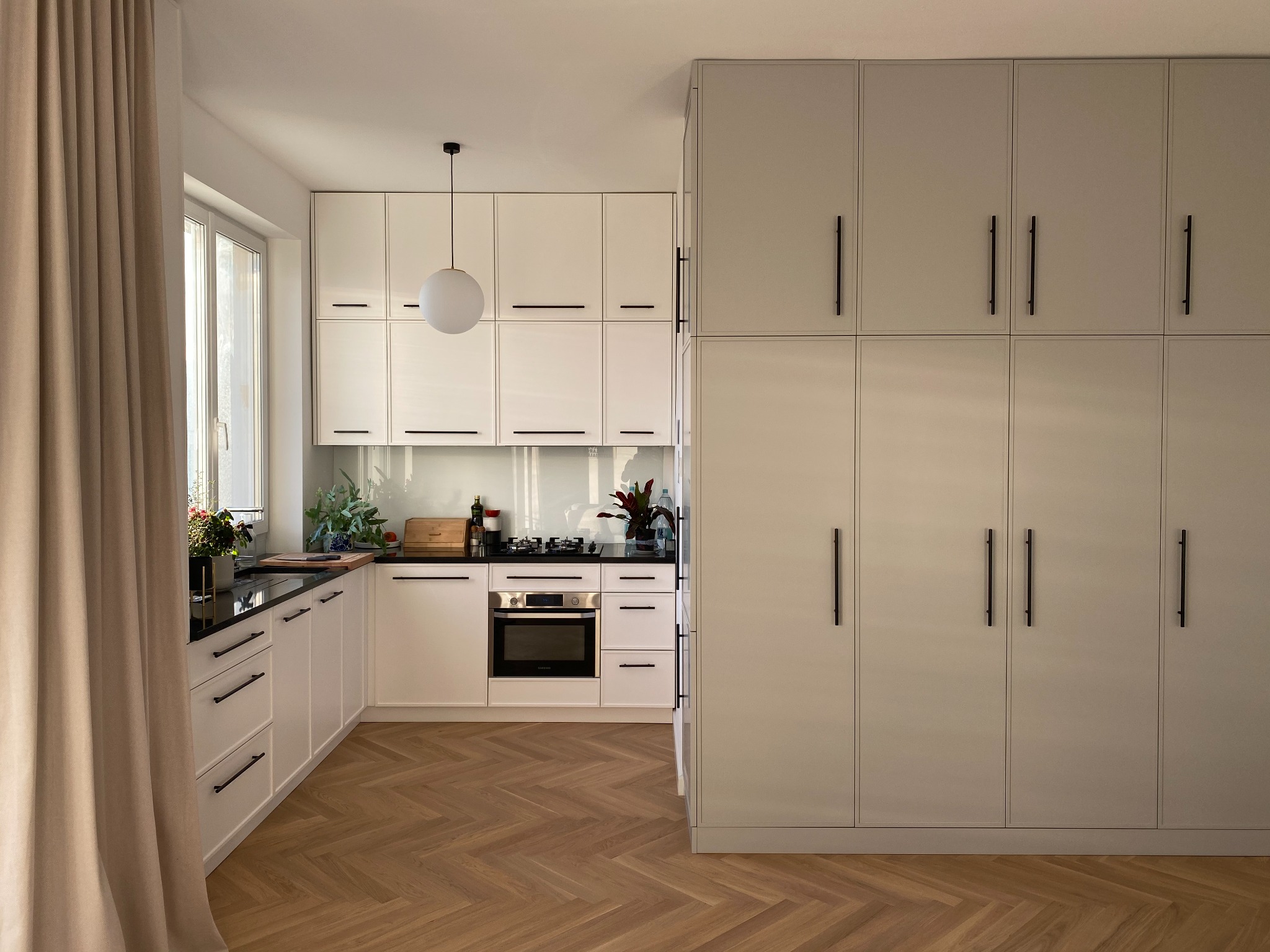 Jasna kuchnia z białymi szafkami z czarnymi uchwytami, czarnym blatem i drewnianą podłogą ułożoną w jodełkę; widoczna zabudowa sięgająca sufitu.