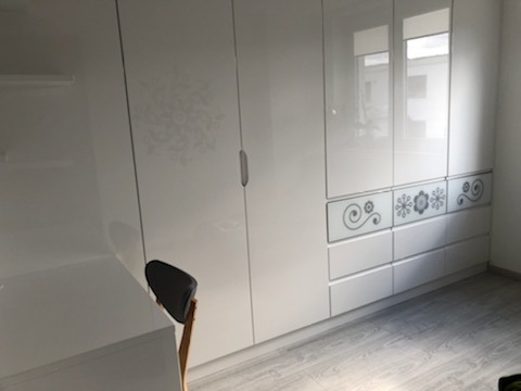 Biała, wysoka szafa z połyskiem i dekoracyjnymi, szarymi wzorami na frontach szuflad, widoczna w pokoju z jasną podłogą i częścią biurka.