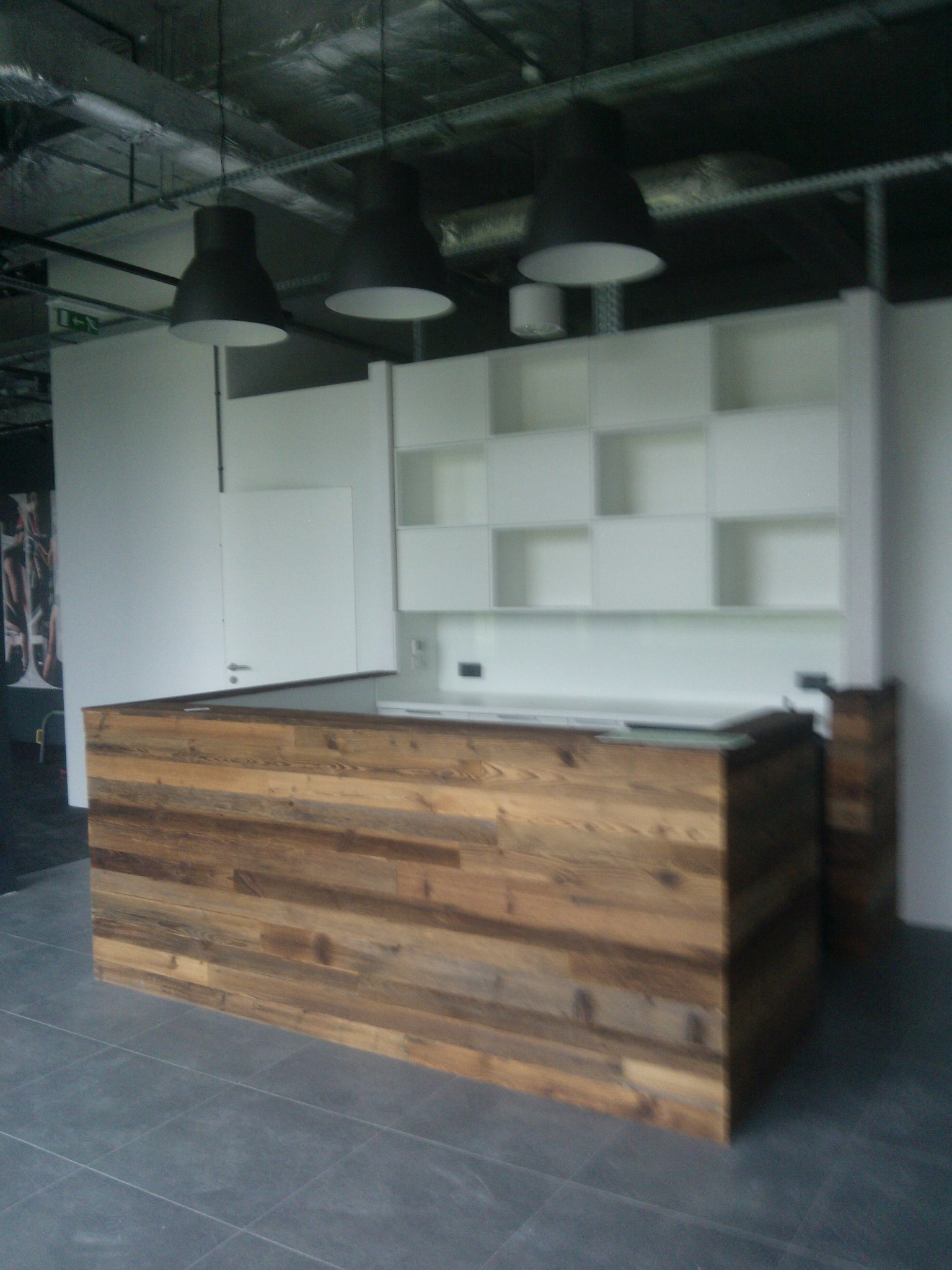 Recepcja z drewnianym frontem z desek o różnej kolorystyce i fakturze, na tle białych szafek z otwartymi półkami i surowego sufitu z widoczną instalacją.