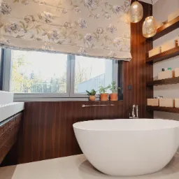 Nowoczesna łazienka z wolnostojącą wanną, drewnianymi akcentami i roletą rzymską w kwiaty, oświetlona wiszącymi lampami.