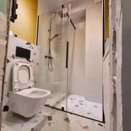 Remont łazienki w trakcie: widoczny wiszący sedes z otwartą klapą, kabina prysznicowa z czarnymi profilami, płytki lastryko na podłodze i ścianie za sedesem, surowy tynk na ścianie obok.