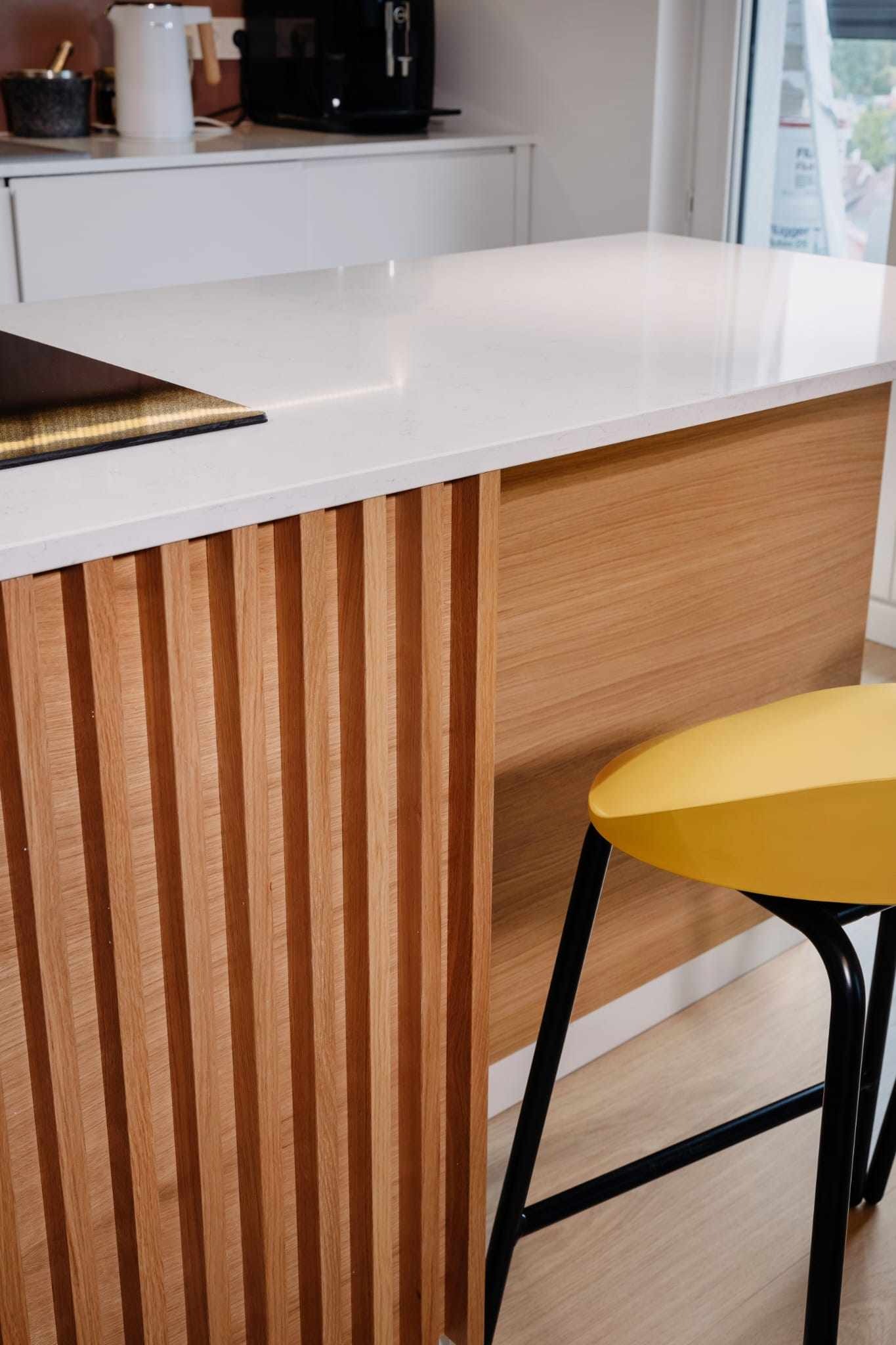 Nowoczesna wyspa kuchenna z białym blatem i drewnianymi panelami dekoracyjnymi; żółty hoker na czarnych nogach.