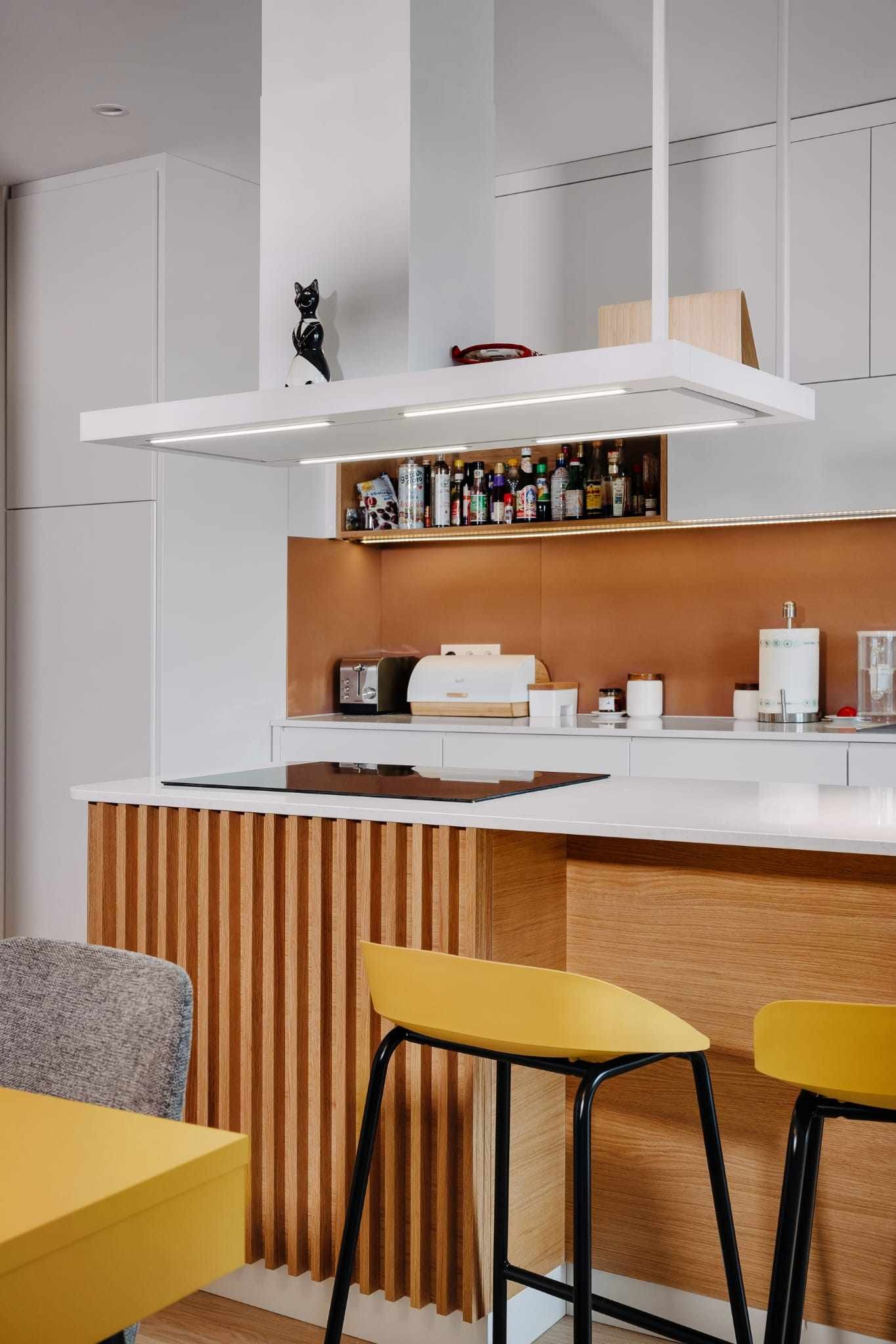 Nowoczesna kuchnia z białymi szafkami, drewnianą wyspą z pionowymi listwami, żółtymi stołkami barowymi i półką z przyprawami podświetloną LED.