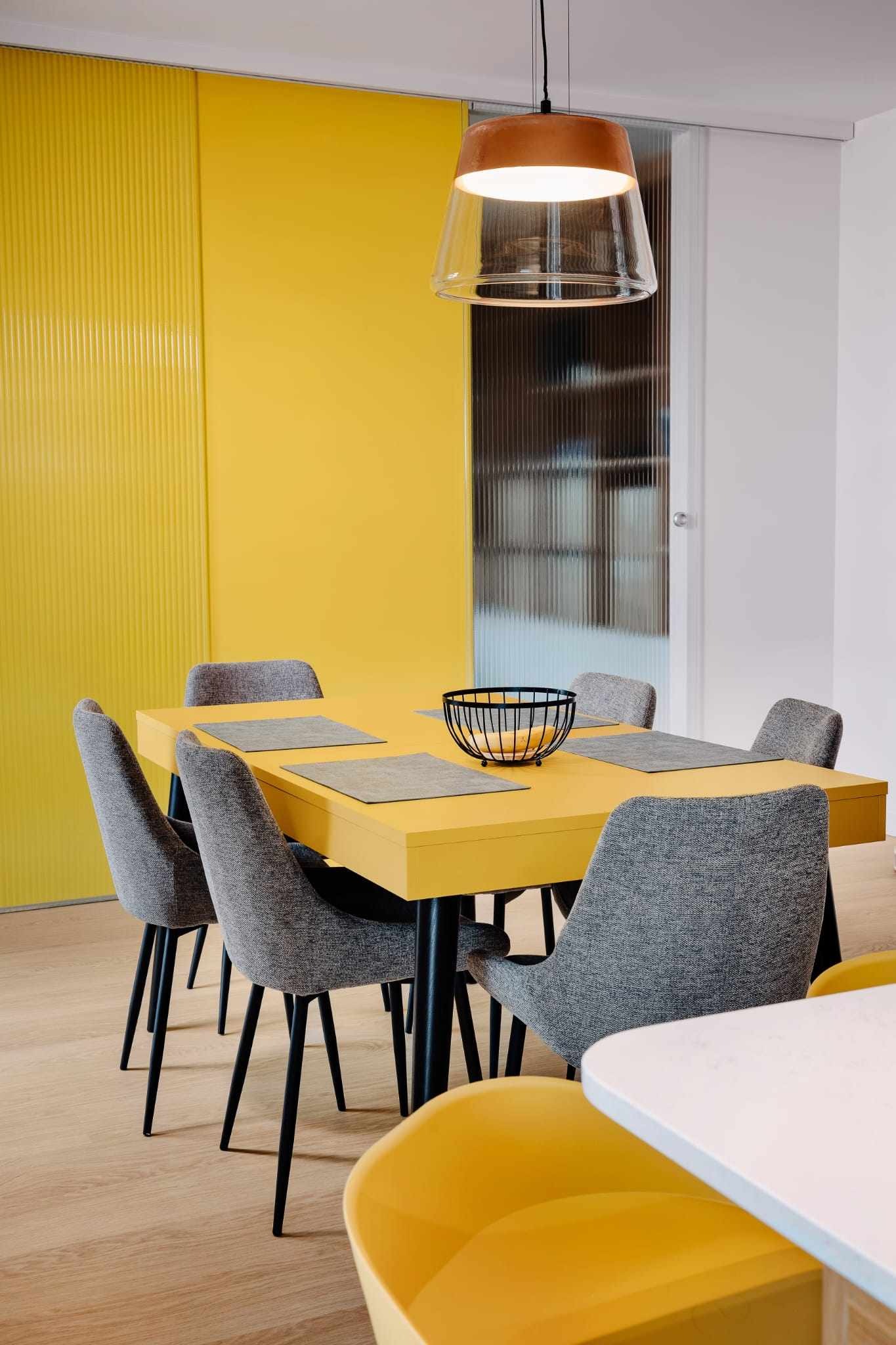 Żółty stół z czarnymi nogami otoczony szarymi krzesłami, na stole szare podkładki i czarny koszyk z owocami, w tle żółta ściana z pionowymi żłobieniami oraz szklane drzwi przesuwne i lampa wisząca...