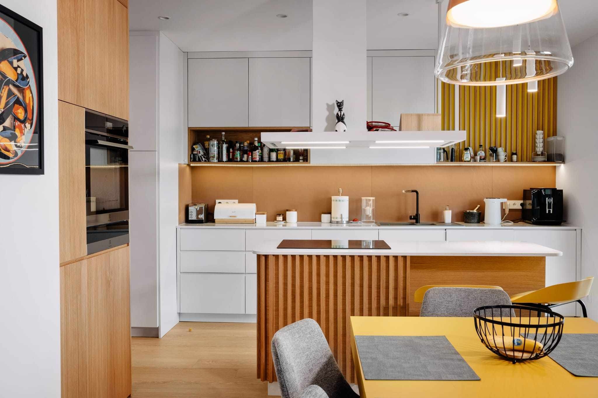 Nowoczesna kuchnia z drewnianymi akcentami: wyspa z lamelami, zabudowa z piekarnikiem i szafkami, żółty stół z szarymi podkładkami, designerska lampa i dekoracyjny obraz na ścianie.