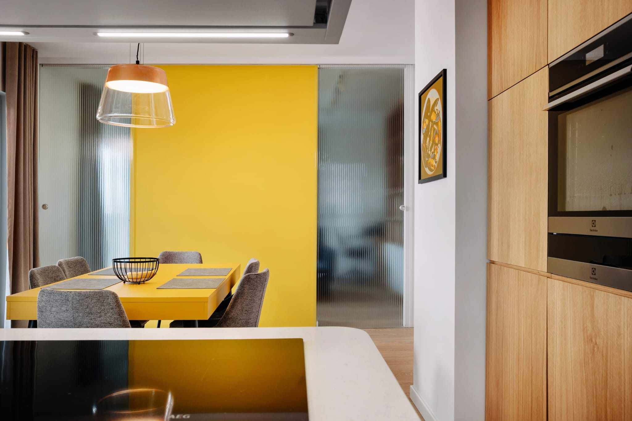 Żółty stół z szarymi krzesłami w nowoczesnej jadalni z drewnianymi szafkami kuchennymi i szklanymi drzwiami przesuwnymi.