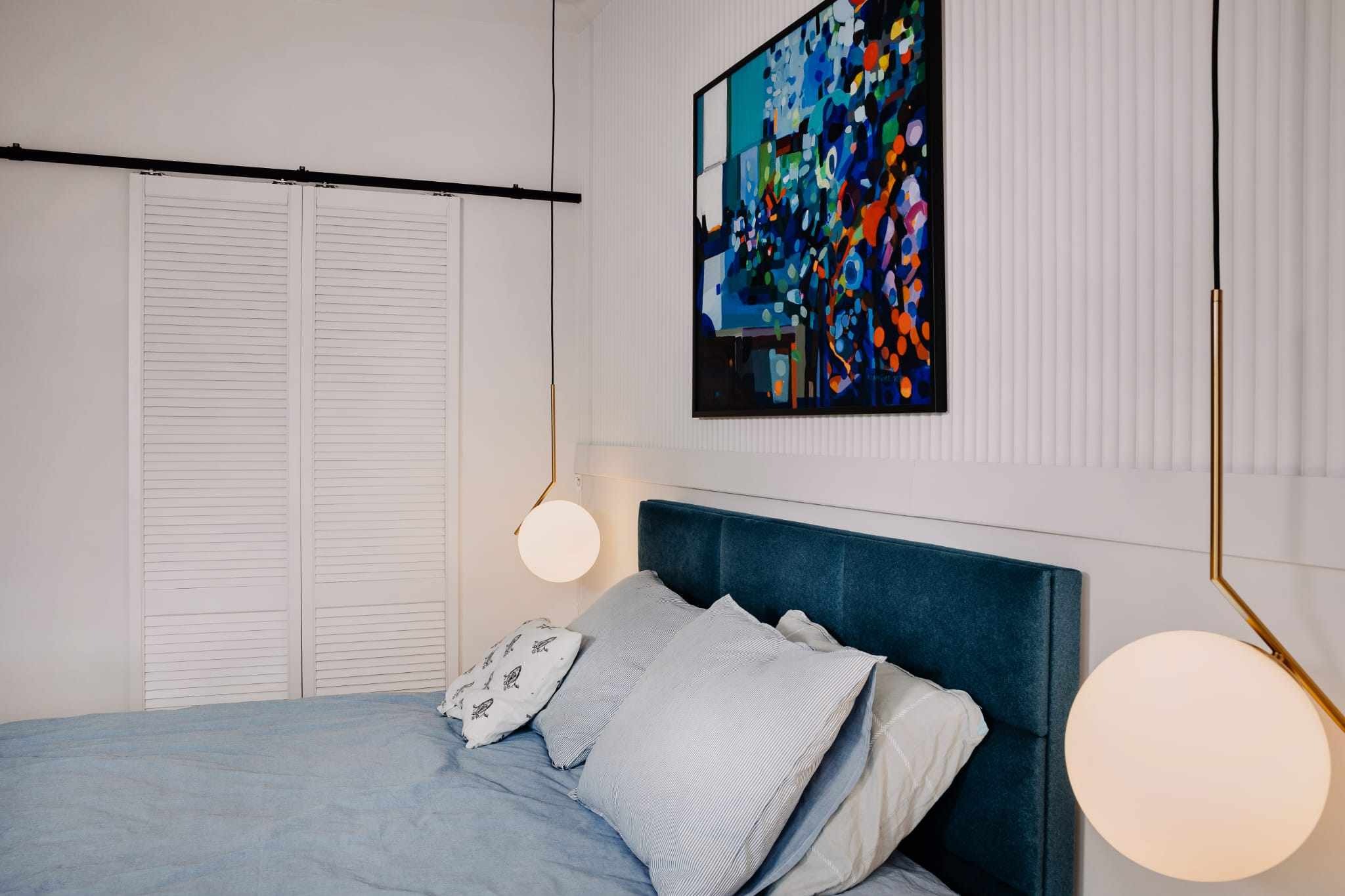 Nowoczesna sypialnia z łóżkiem w niebieskich odcieniach, ozdobiona abstrakcyjnym obrazem i kulistymi lampami wiszącymi na złotych prętach, z białymi, żaluzjowymi drzwiami w tle.