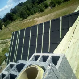 Widok z g&oacute;ry na konstrukcję dachu w trakcie budowy, z widoczną membraną dachową, łatami, kontrłatami oraz kominem z szarego pustaka z osadzoną rurą, na tle zieleni i błękitnego nieba.