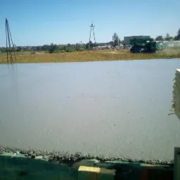 Świeżo wylana, gładka płyta fundamentowa z widocznym szalunkiem drewnianym i elementami zbrojenia wystającymi z betonu. W tle krajobraz polny i maszyna budowlana.