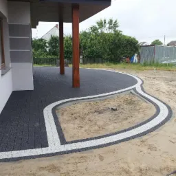 Ułożona, ciemnoszara nawierzchnia z jasnymi obrzeżami, tworząca ścieżkę wokół piaskowego klombu przy budynku z drewnianymi kolumnami.