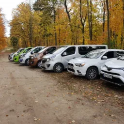 Siedem samochodów osobowych (w tym jeden bus) różnych marek ustawionych w rzędzie na leśnym parkingu w jesiennej scenerii. Widoczne tablice rejestracyjne.