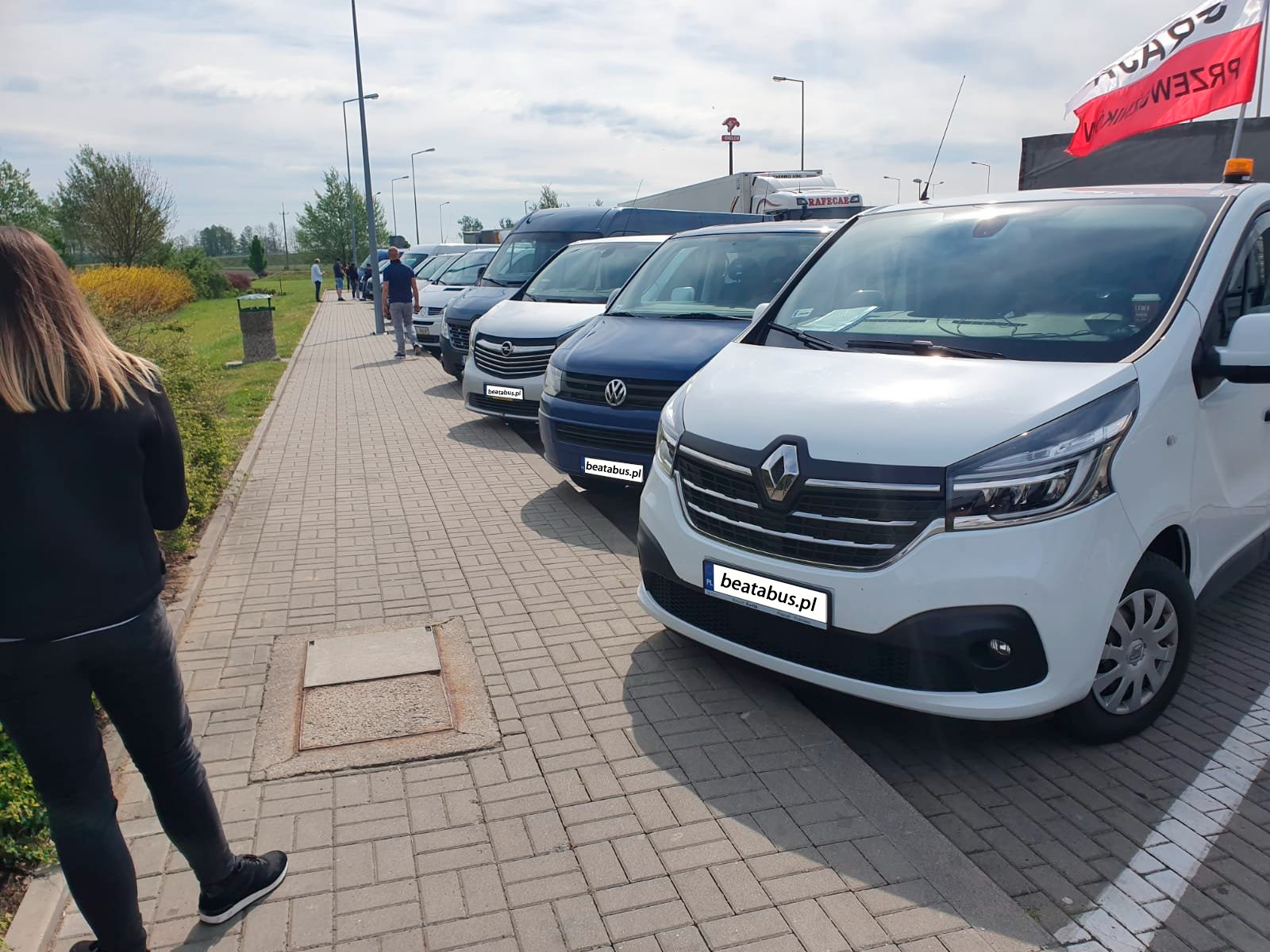 Rząd zaparkowanych busów transportowych, z widocznym logo firmy na tablicach rejestracyjnych i flaga Polski na jednym z pojazdów, pasażerowie w tle.