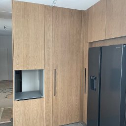 Ar_inwestycje - Montaż zabudowy kuchennej z drewnianymi frontami i czarną lodówką side-by-side w Kielcach, widoczny etap prac wykończeniowych z zabezpieczoną podłogą.