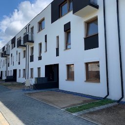 AR - Inwestycje Fatyga Rafał - Nowoczesny szeregowiec z białymi ścianami i czarnymi elementami dekoracyjnymi, widoczne okna z drewnianą ramą i balkony z czarną balustradą, teren wokół budynku jeszcze w trakcie zagospodarowania...