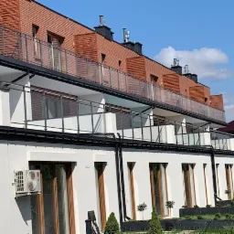 Nowoczesny budynek wielorodzinny z balkonami z przezroczystymi balustradami i elewacją w kolorze białym, z klimatyzatorem na ścianie i dekoracyjnymi drzewkami w donicach przed budynkiem.