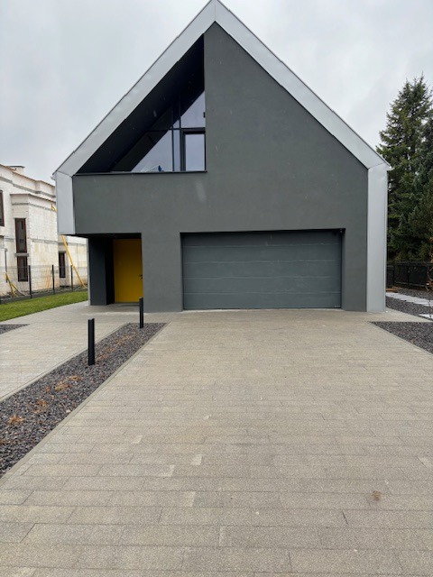 Nowoczesny dom murowany z garażem, szara elewacja, żółte drzwi wejściowe, trapezowe okno na poddaszu, podjazd wyłożony kostką brukową.