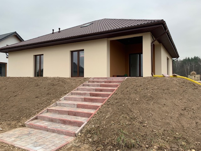 Parterowy dom z beżową elewacją i brązowym dachem, widoczny z dołu schodów prowadzących na posesję, z ziemią do wyrównania wokół.