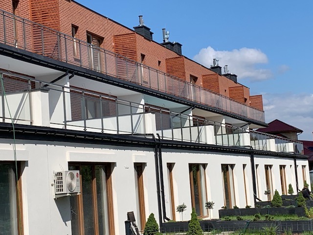 Nowoczesny budynek wielorodzinny z balkonami z przezroczystymi balustradami i elewacją w kolorze białym, z klimatyzatorem na ścianie i dekoracyjnymi drzewkami w donicach przed budynkiem.