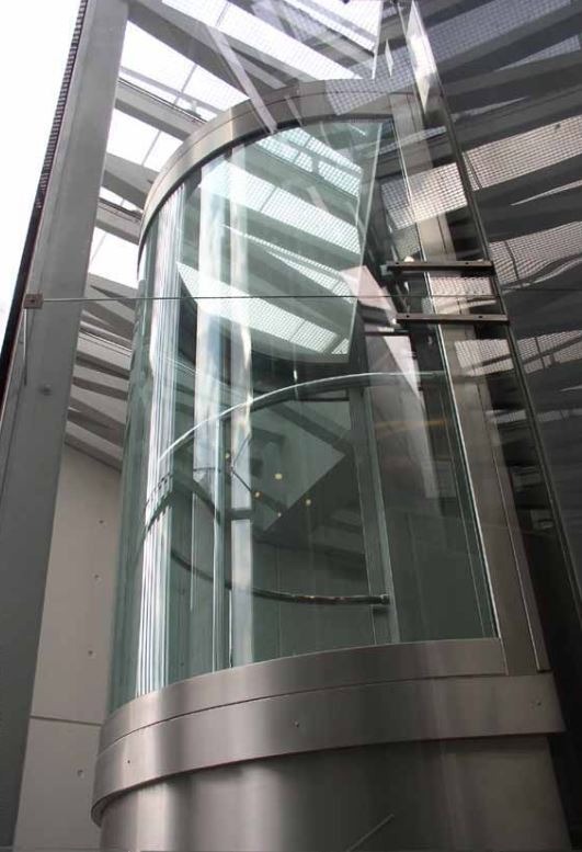 Szklana, cylindryczna winda panoramiczna w nowoczesnym budynku biurowym z widocznymi elementami konstrukcyjnymi i metalowymi wykończeniami.