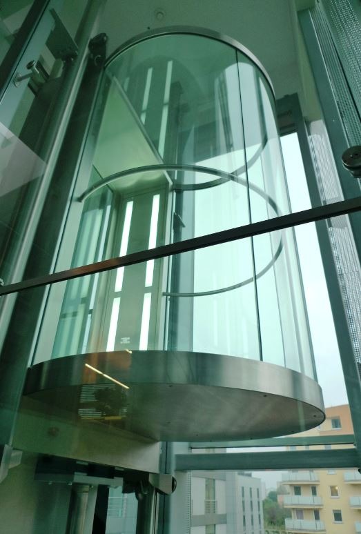 Szklana panoramiczna kabina osobowa w szybie windowym, widok z dołu na górę, przez szybę widoczne budynki mieszkalne.