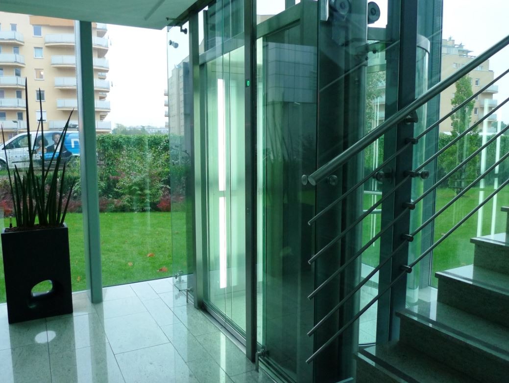 Szklana kabina dźwigu osobowego z oświetleniem wewnętrznym, widoczna z holu budynku z przeszkloną ścianą, za którą rozciąga się trawnik i zabudowa miejska.