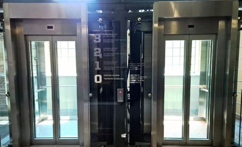 Trzy metalowe windy z przeszklonymi drzwiami, środkowa z numerami 3, 2, 1, 0 i napisem 'Wystawa Czasowa' wewnątrz budynku.
