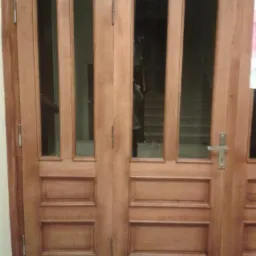 Dwuskrzydłowe drewniane drzwi z przeszkleniami, widoczny fragment klatki schodowej w tle, metalowy uchwyt i zamek.