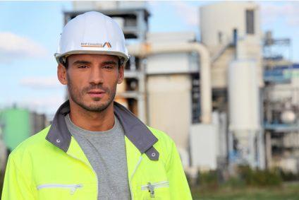 Mężczyzna w kasku i jaskrawej kurtce na tle instalacji przemysłowej. Kask z logo firmy. Ujęcie portretowe, skupione na twarzy.