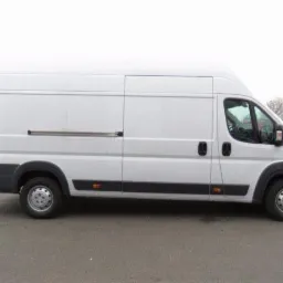 Biały, wysoki bus dostawczy Fiat Ducato z przesuwnymi drzwiami bocznymi, zaparkowany na szarym asfalcie, widok z boku.