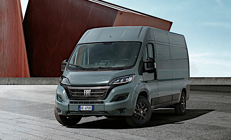 Szary bus marki Fiat Ducato, z czarnymi felgami, zaparkowany na betonowym placu na tle nowoczesnej, rdzawoczerwonej konstrukcji architektonicznej.